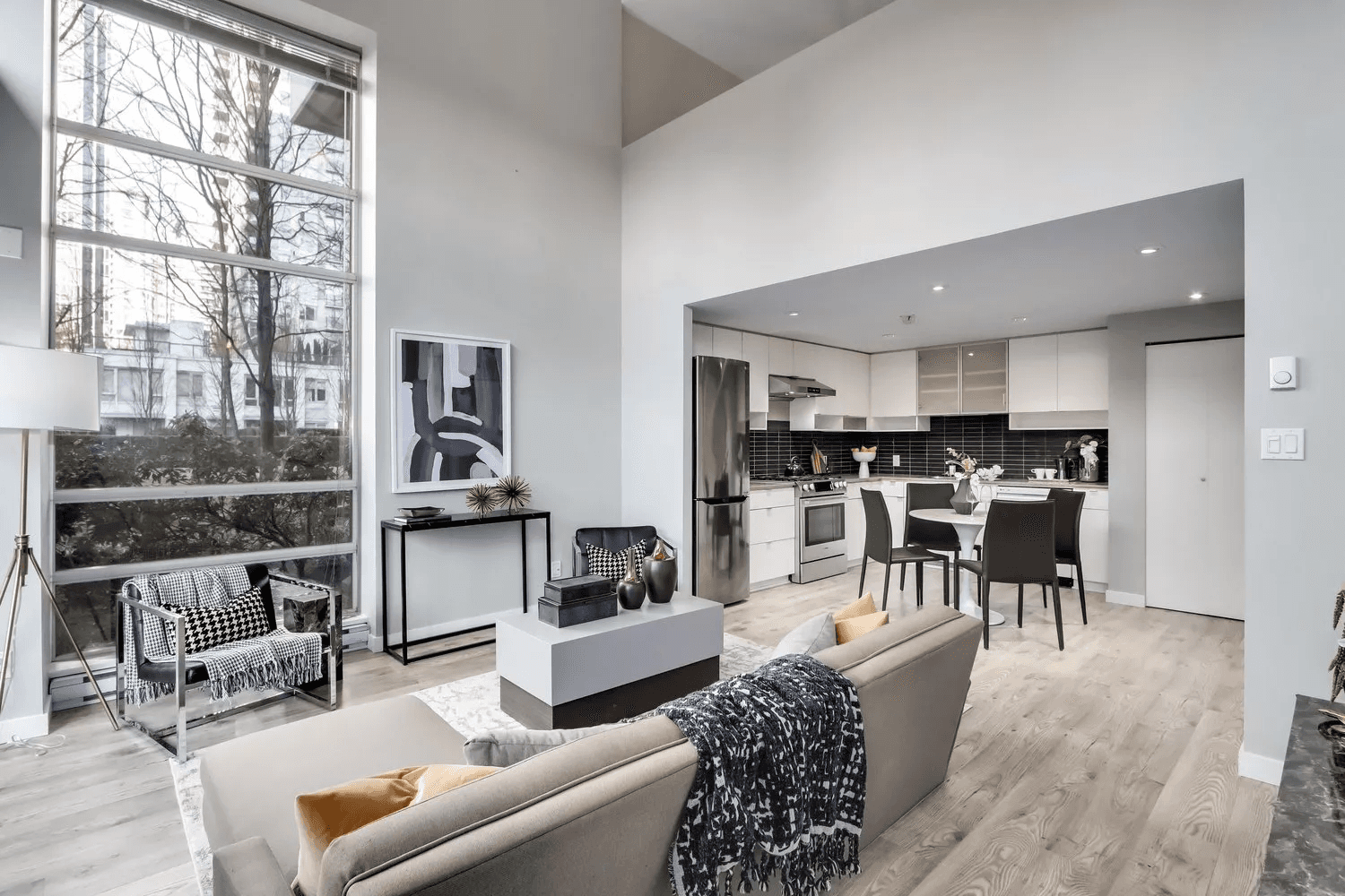 Modern living room with high ceilings, large windows, and an open kitchen, featuring minimalistic furnishings.