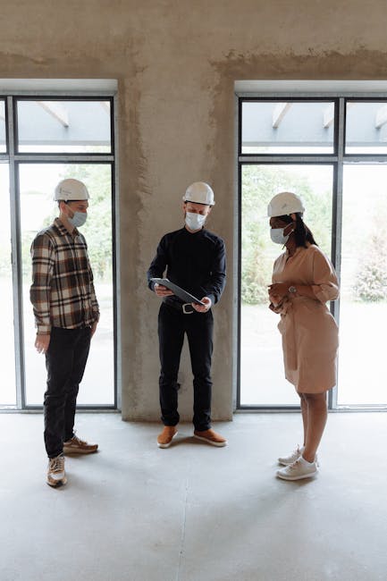 Three people in masks and hard hats discuss real estate indoors with natural lighting.