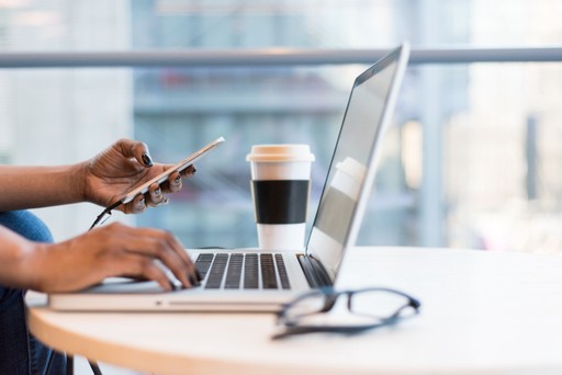 A person types on a laptop and holds a smartphone, with a coffee cup nearby, in a bright workspace.