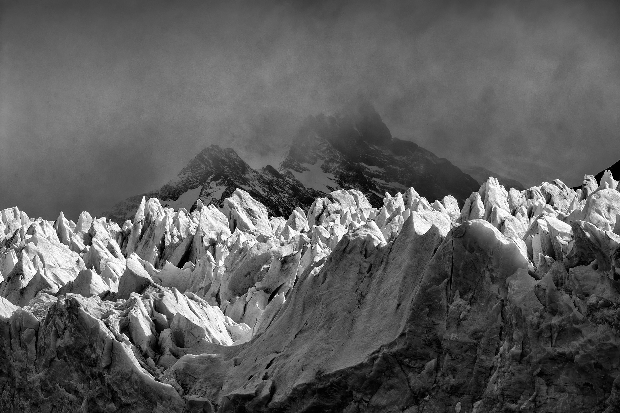 Sharp ice peaks of a glacier reach upward beneath storm clouds, in high-contrast black and white