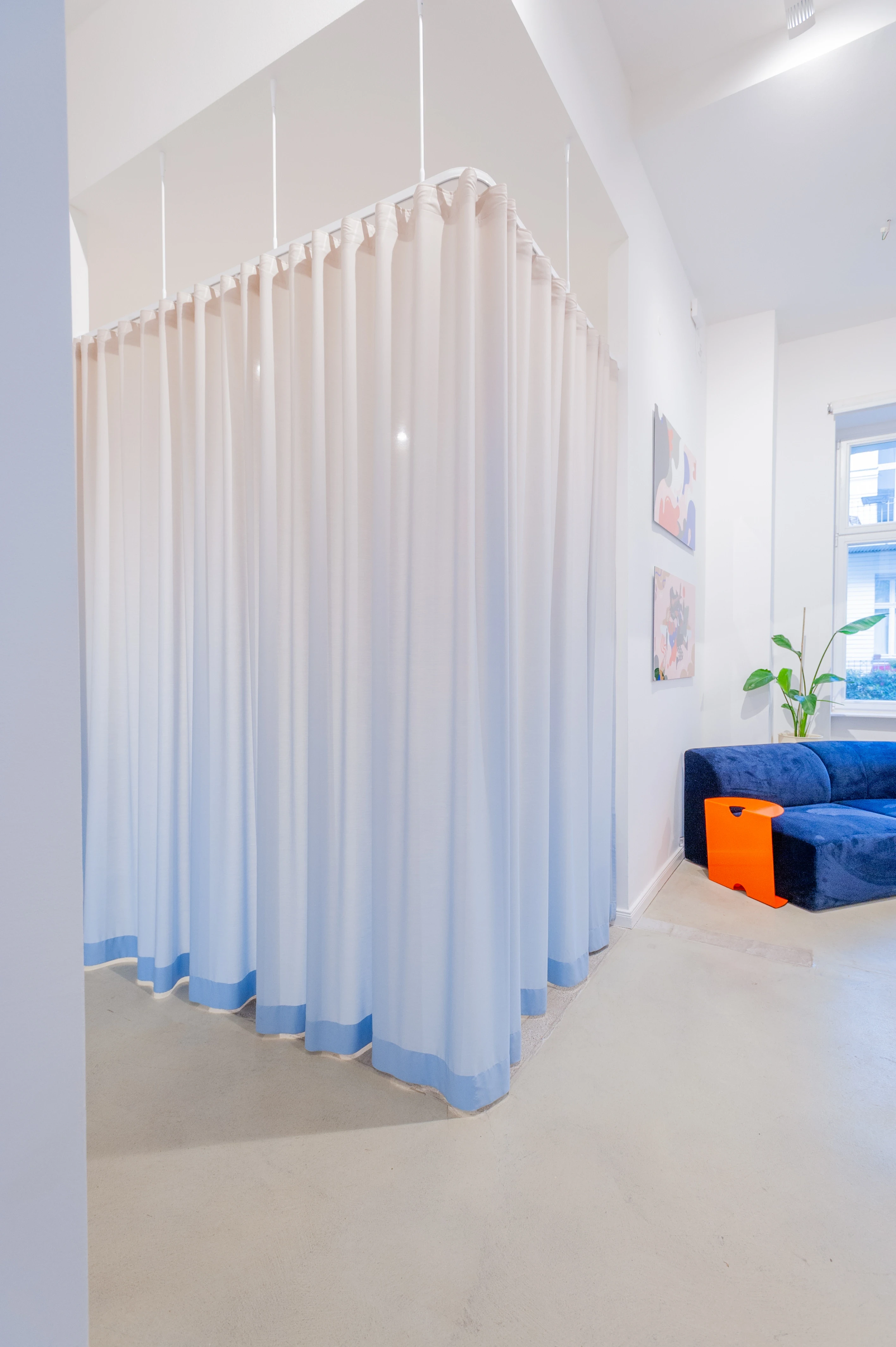 White curtain cube at BATTLE ROYAL hanging from a slim ceiling-mounted rail, with light blue trim at the hem, next to a navy velvet sofa with an orange side table and small artworks.