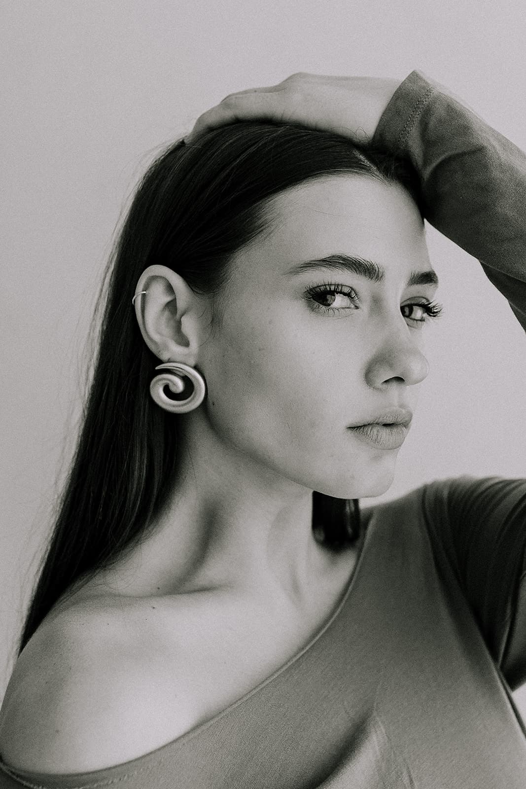 A model gently rests her hand on her head, displaying gold spiral earrings and a neutral expression, in a natural light studio in Shreveport.