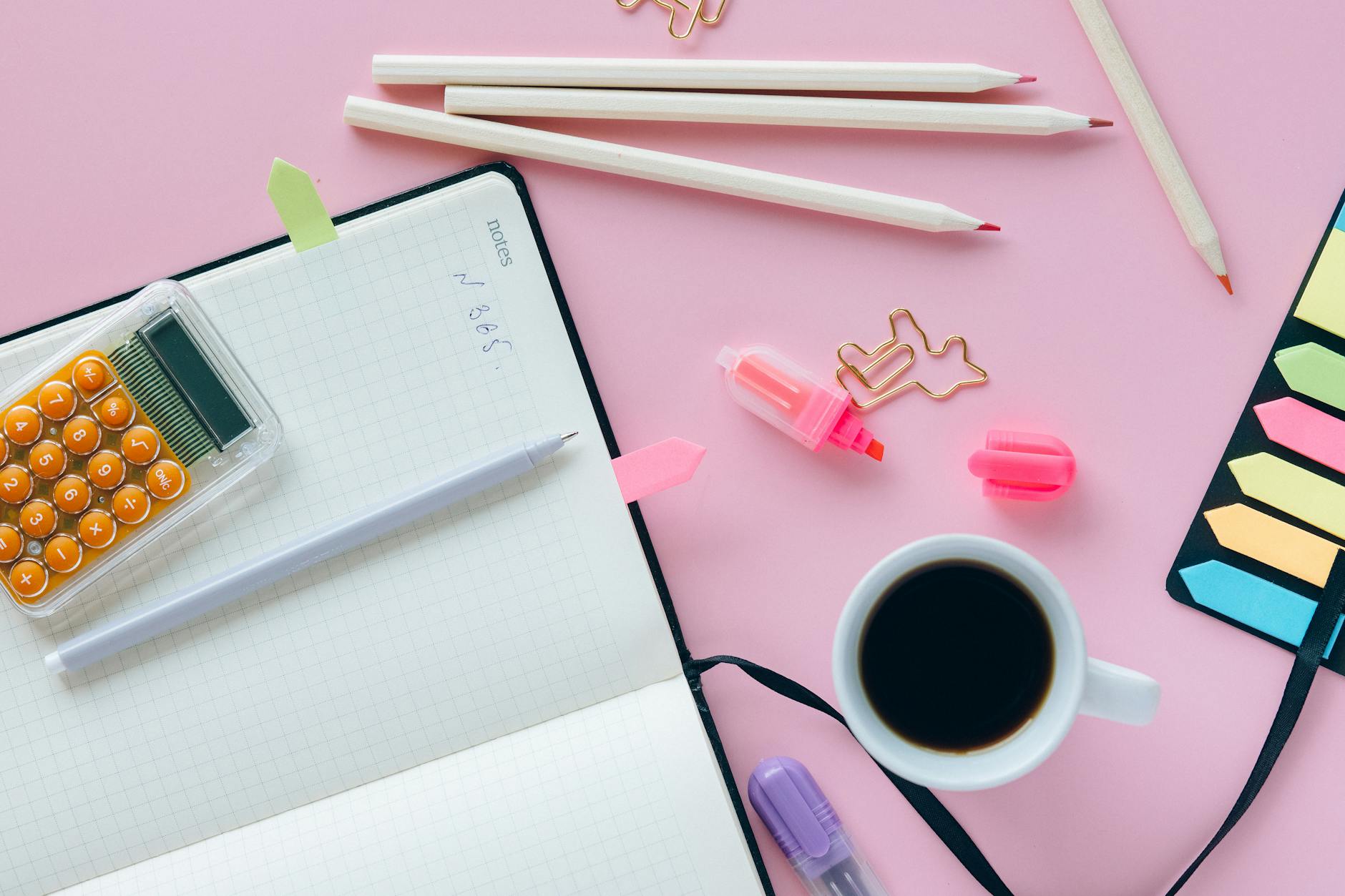 Flatlay of work essentials on a pink background featuring coffee and stationery.