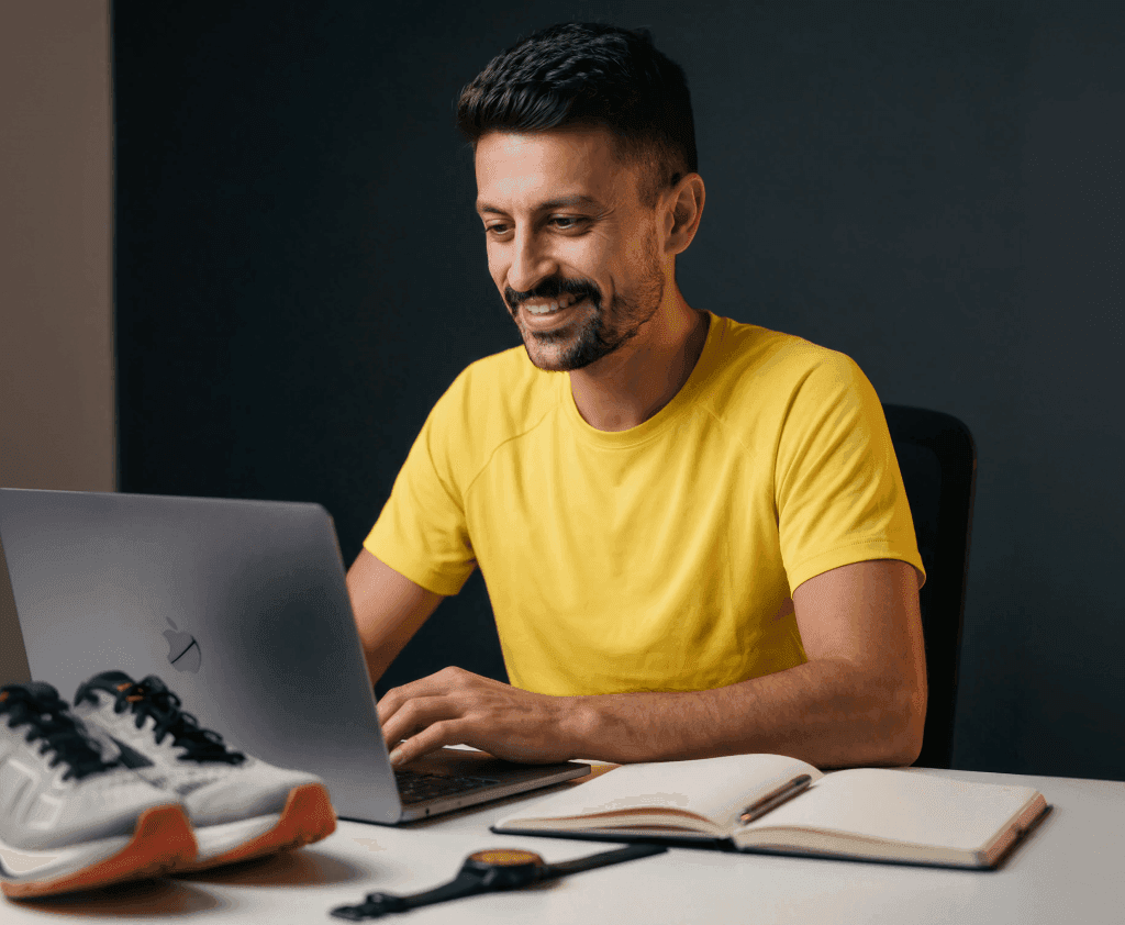Treinador de corrida de rua sentado, sorrindo e olhando pra tela do notebook em cima de uma mesa com: caderno, caneta, relógio e tênis de corrida