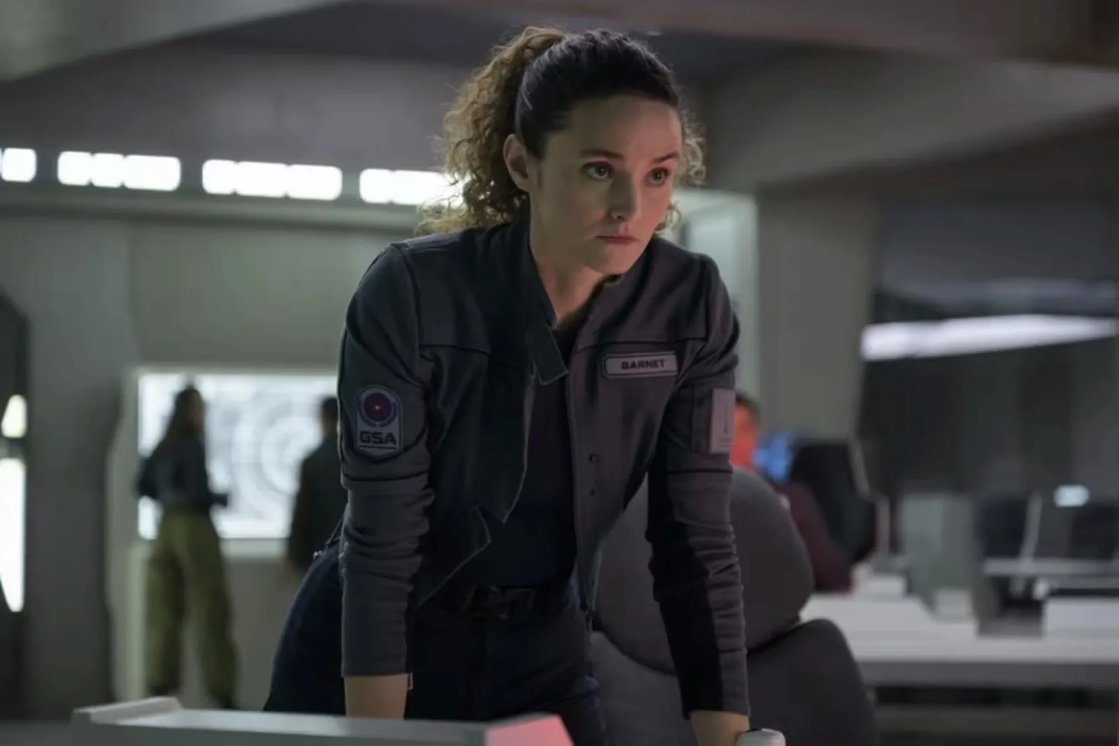 A woman with curly hair, wearing a dark uniform with a space organization patch, leans over a console in a futuristic control room.