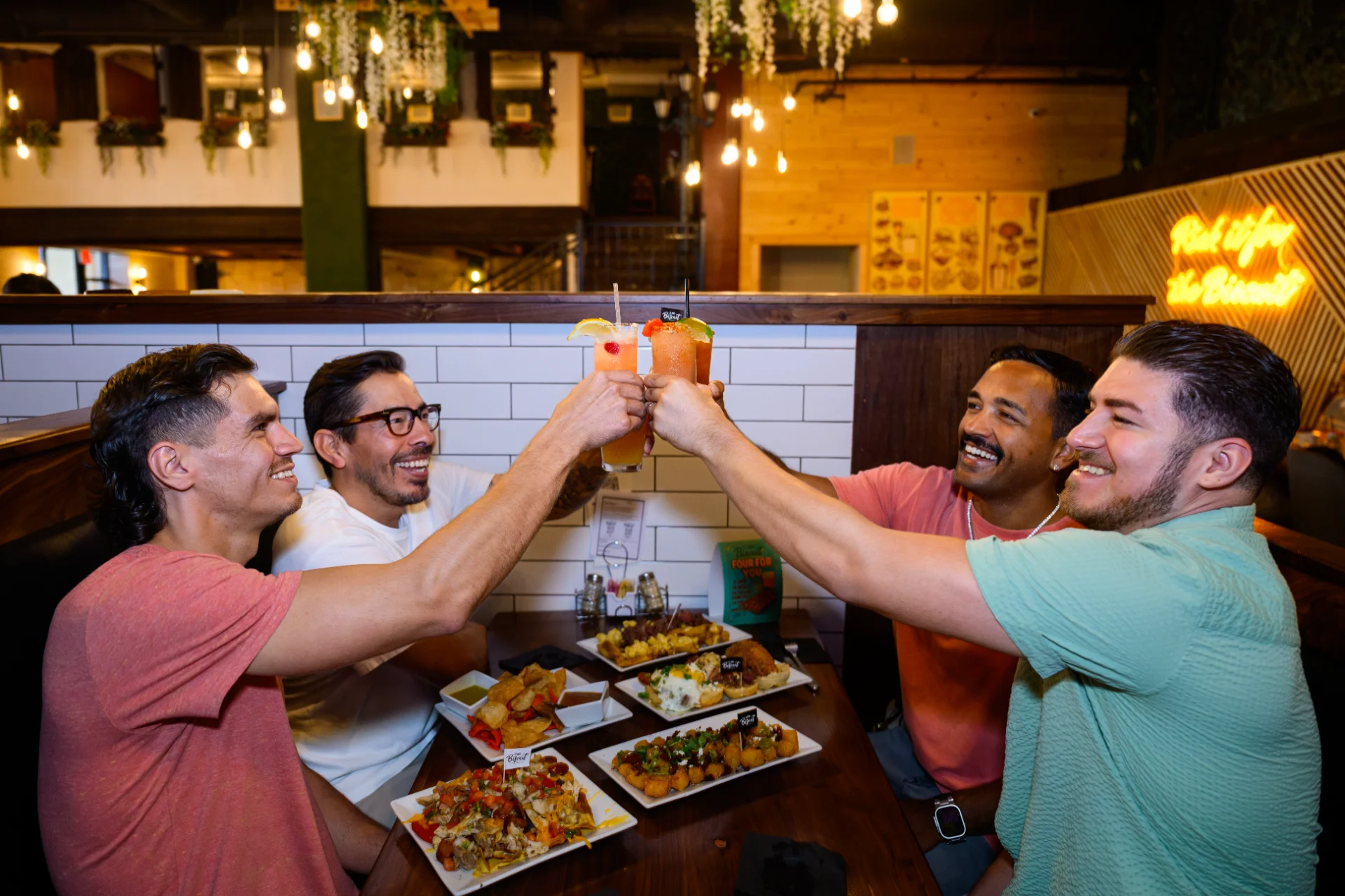 Four friends toasting cocktails over a shared appetizer spread at Alamo Biscuit