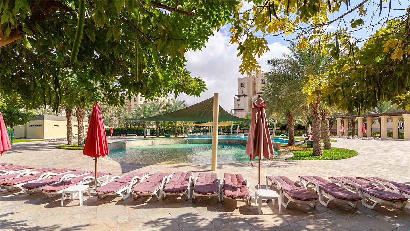 Relaxing poolside in Remraam Dubai with maroon loungers, red umbrellas, and lush greenery in a resort-style setting.
