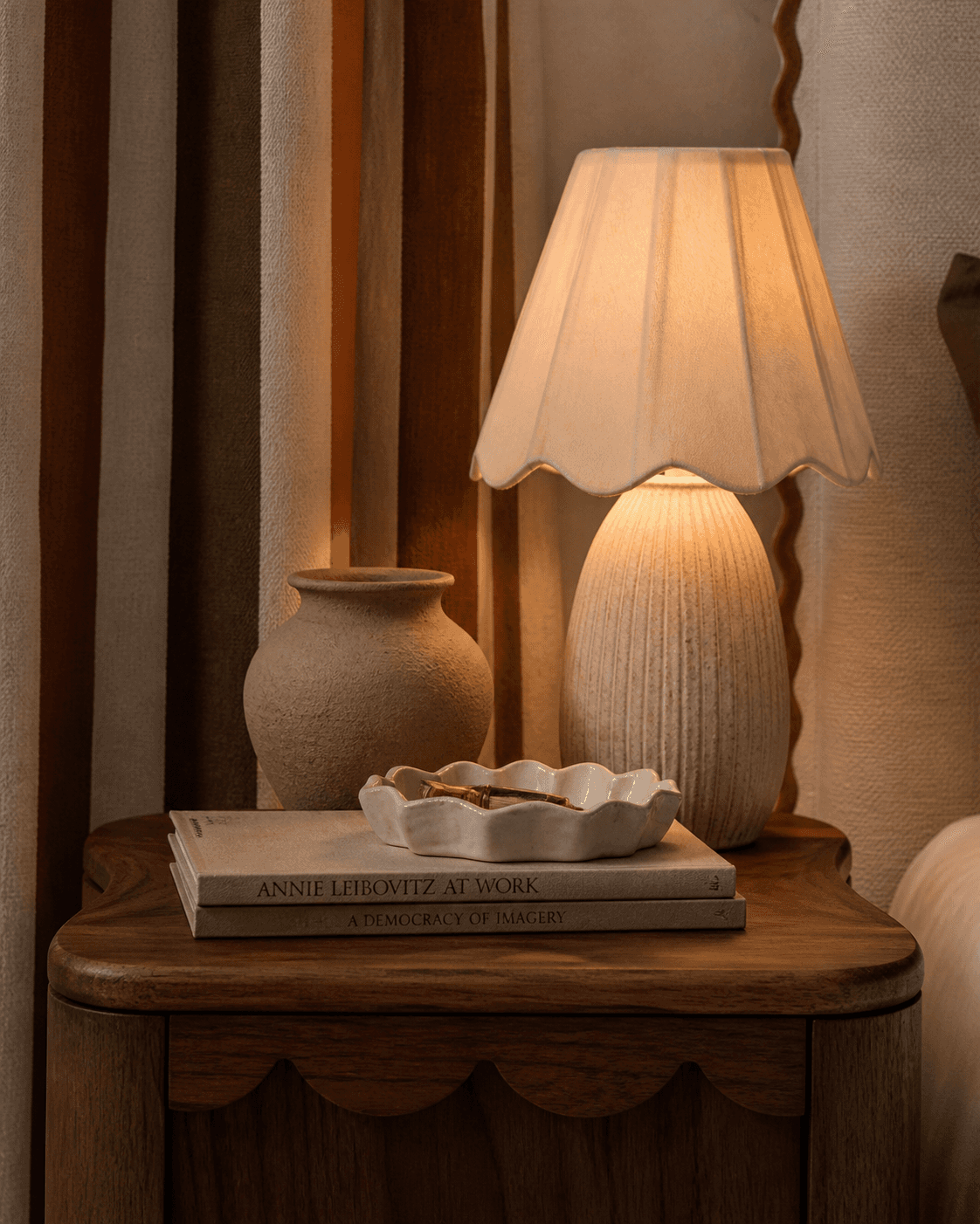 Refined circuscore bedside table vignette featuring a warm walnut nightstand with soft rounded edges and subtle scalloped detailing, styled with a ceramic table lamp with a pleated scalloped shade, neutral stacked books, a matte clay vase, and a scalloped ceramic tray. Behind the table, painterly striped drapery in ochre, clay, ivory, and deep olive creates a soft, theatrical backdrop. The scene is softly lit with warm ambient light, highlighting tactile materials like linen, plaster, and wood in a calm, minimal, high-end interior design style.