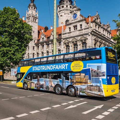 「STADTRUNDFAHRT」と都市の画像が描かれた青と黄色の二階建てバスが、都市の街路にある歴史的な建物の近くに停まっている。