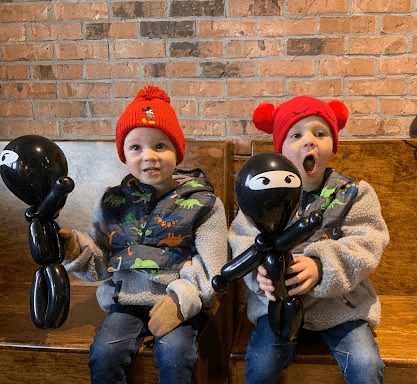Two young boys wearing winter hats and holding black ninja-shaped balloon figures, sitting on a wooden bench against a brick wall
