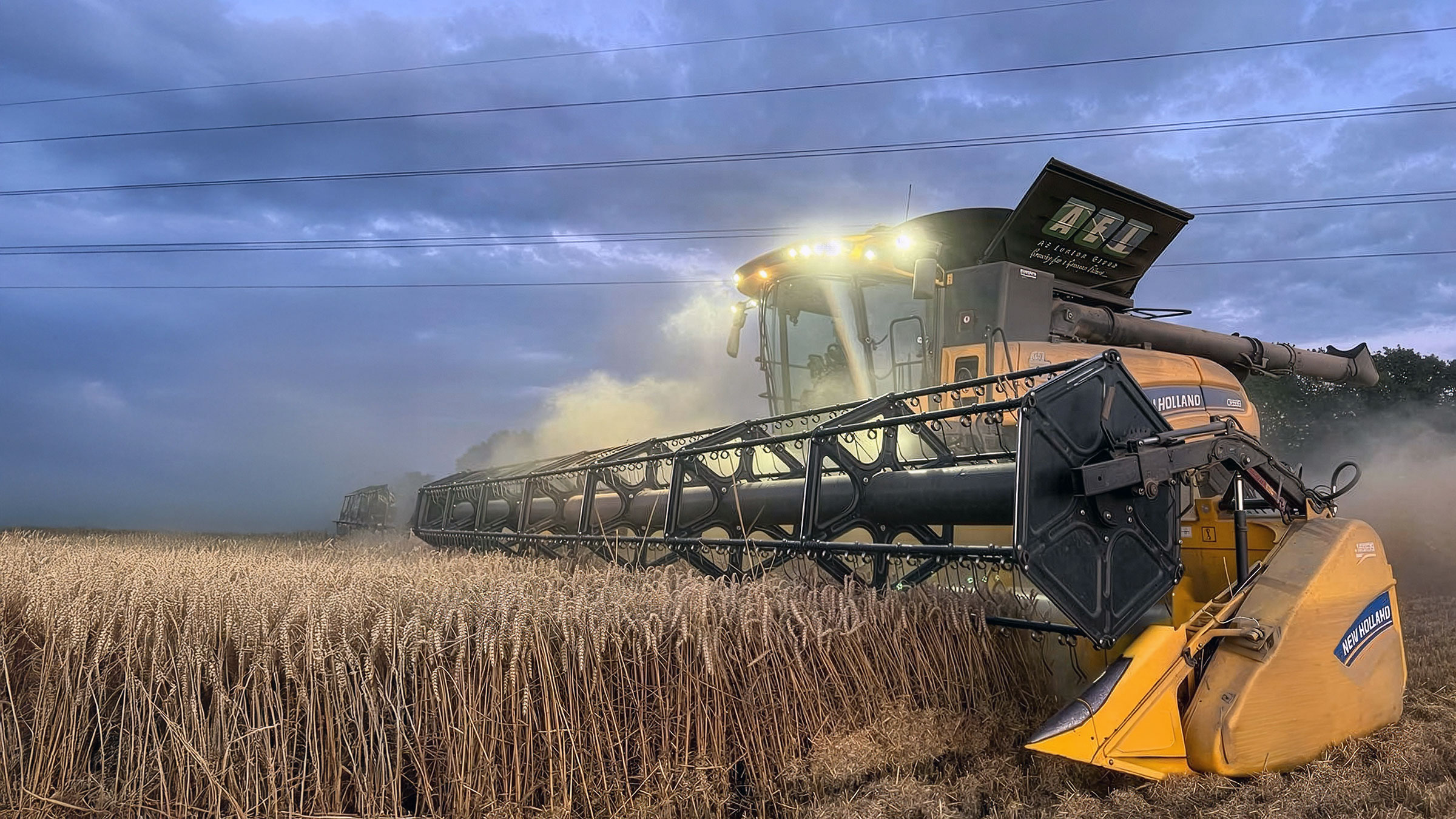 New Holland combine harvester with lights on cutting wheat field at dusk under stormy blue sky