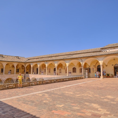 A courtyard with a rectangular building featuring arches and a tiled roof, with people walking and standing on the paved ground.