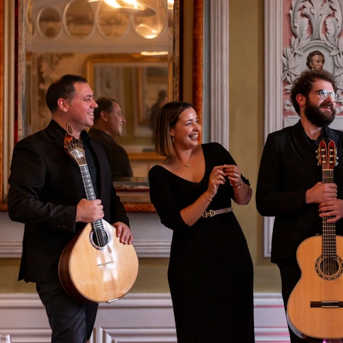 Three people in formal attire stand indoors, two holding guitars and one in the middle smiling. The room has ornate decorations and mirrors.