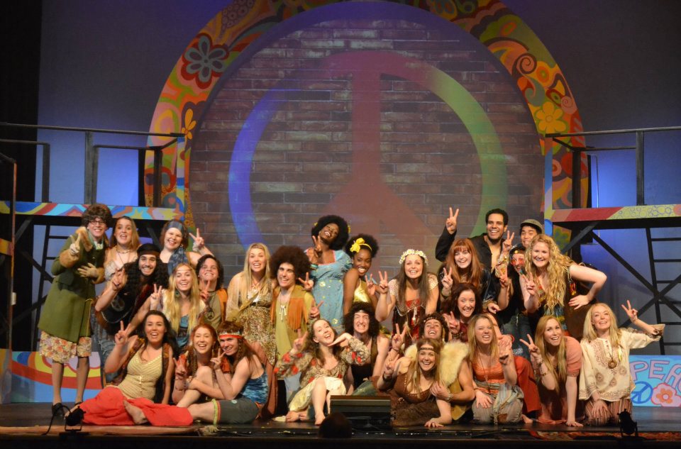 Large group of smiling performers posing on a brightly lit stage in colorful costumes after a musical theatre production.