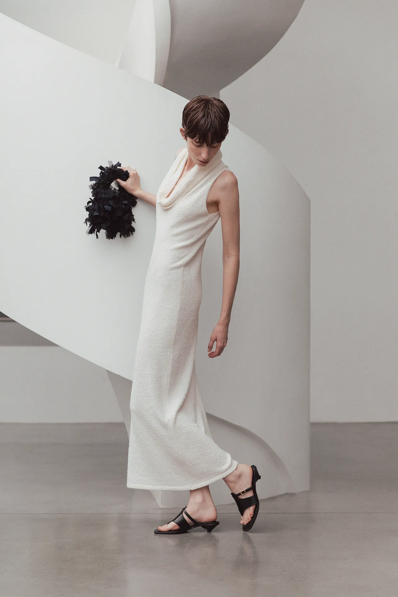 Model wearing the Aida Barni SS26 collection white textured sleeveless knit dress with a draped cowl neckline, holding a dark fringed bag.