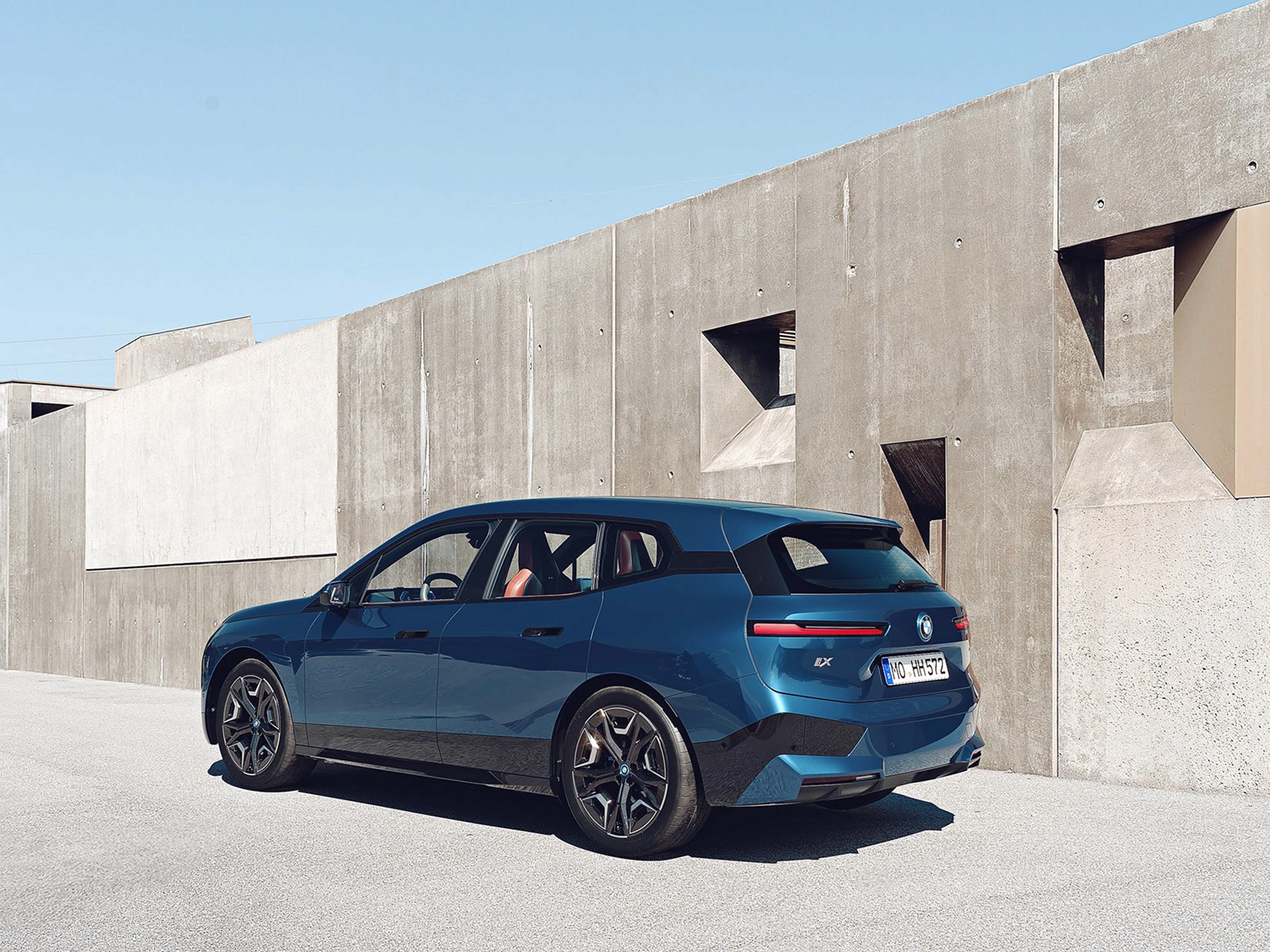 Vista tre quarti posteriore di BMW iX con carrozzeria blu metallizzata in ambiente architettonico moderno e minimalista in cemento chiaro.