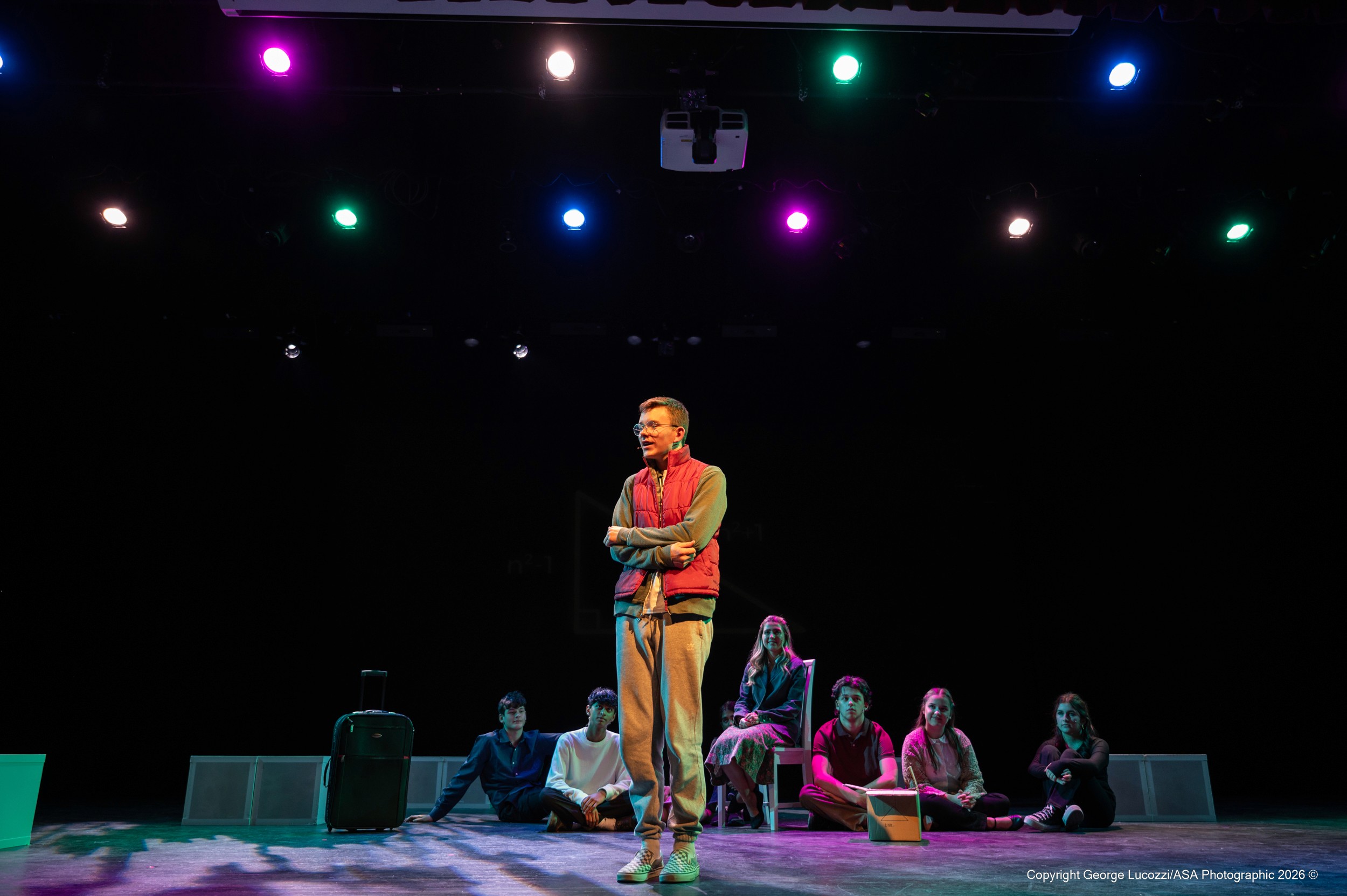 During the Maths Appendix, Chris stands downstage in a bright followspot, smiling as saturated pink, green, blue, and teal stage washes fade dynamically behind him, framing the cast in celebratory color transitions.
