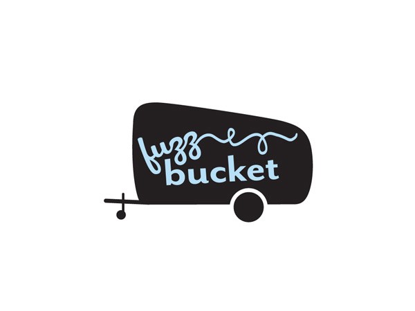 A white model Airstream trailer parked on a light surface, featuring a sign that encourages knitting. Miniature figures nearby engage with the scene, while a chalkboard lists knitting lesson times. The trailer has a vibrant design, enhancing its inviting atmosphere for both knitters and those new to the craft.