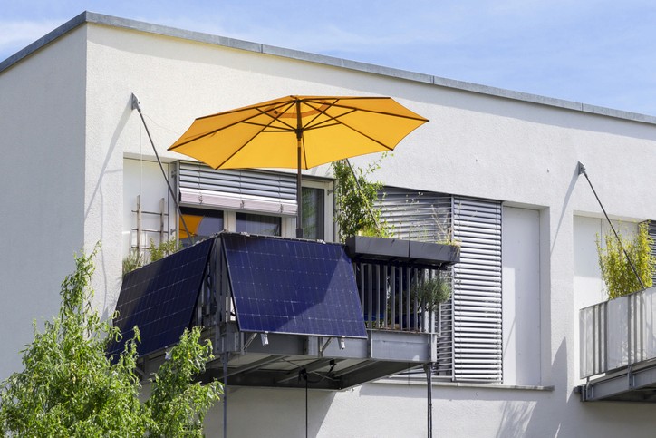 Ein Balkon mit zwei installierten Solarpaneelen, umgeben von Grünpflanzen und einem modernen Gebäude im Hintergrund.