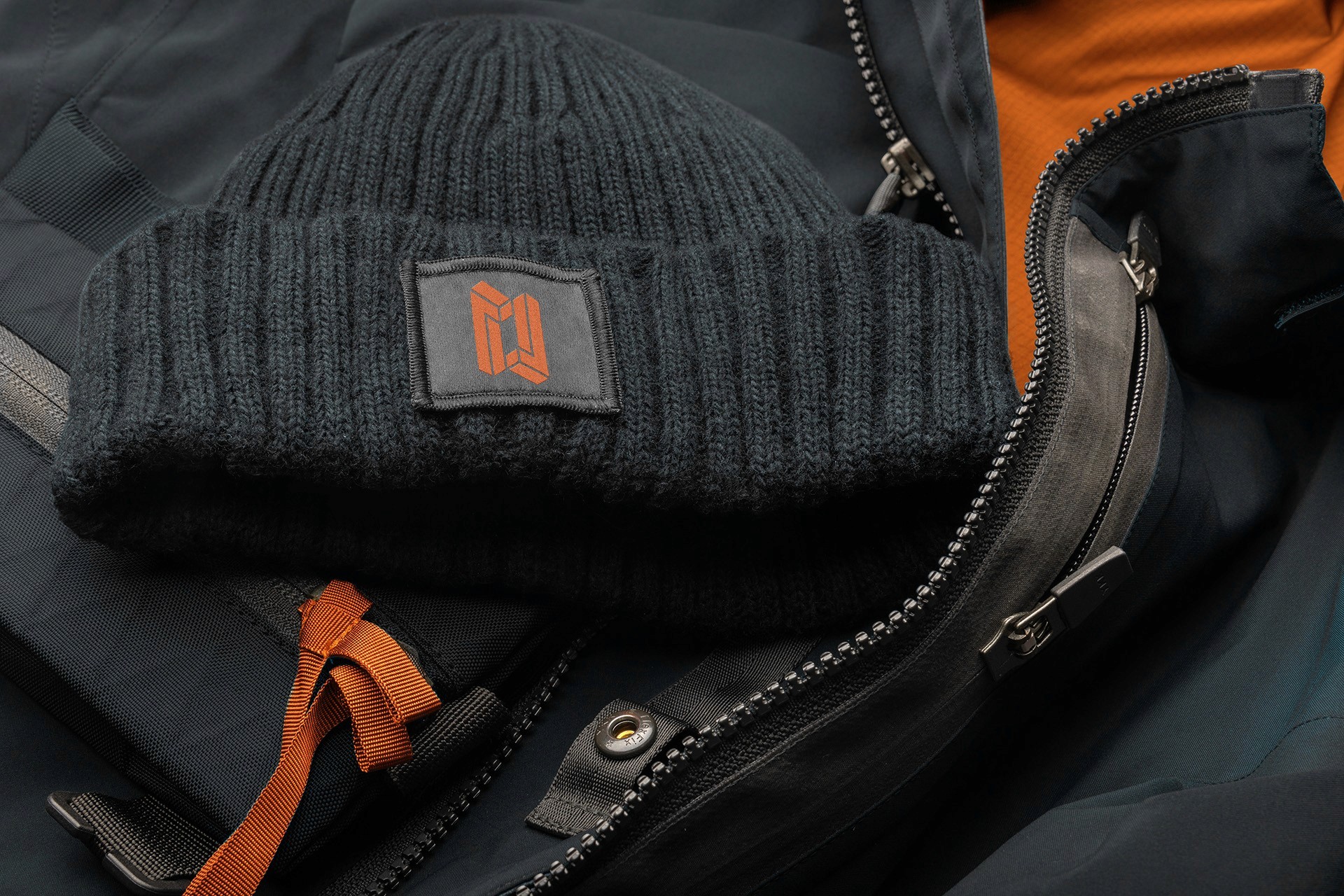 Schwarze Strickmütze mit orangefarbenem Logo-Patch, drapiert auf einer schwarzen Jacke mit orangefarbenen Akzenten und Reißverschlussdetail.
