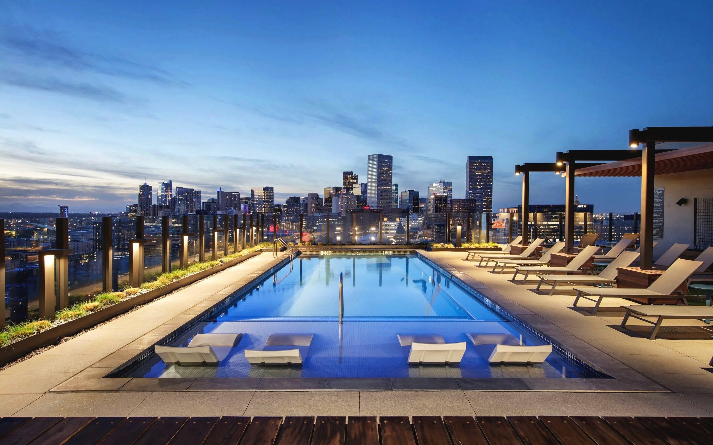 Rooftop pool and lounge chairs overlook city skyline at dusk.