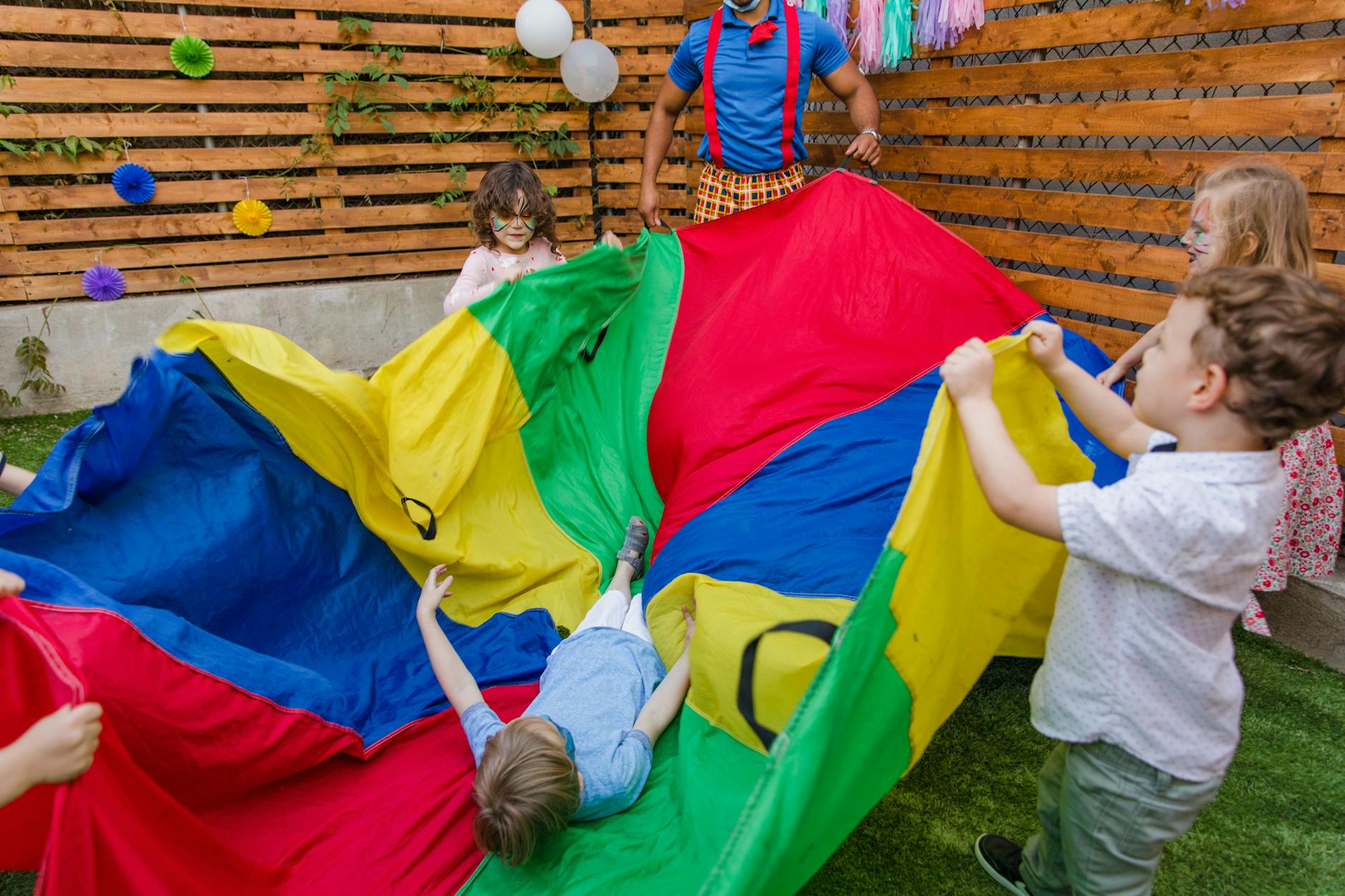 Elementary students laughing while working together to balance a large colorful parachute in a bright gymnasium.