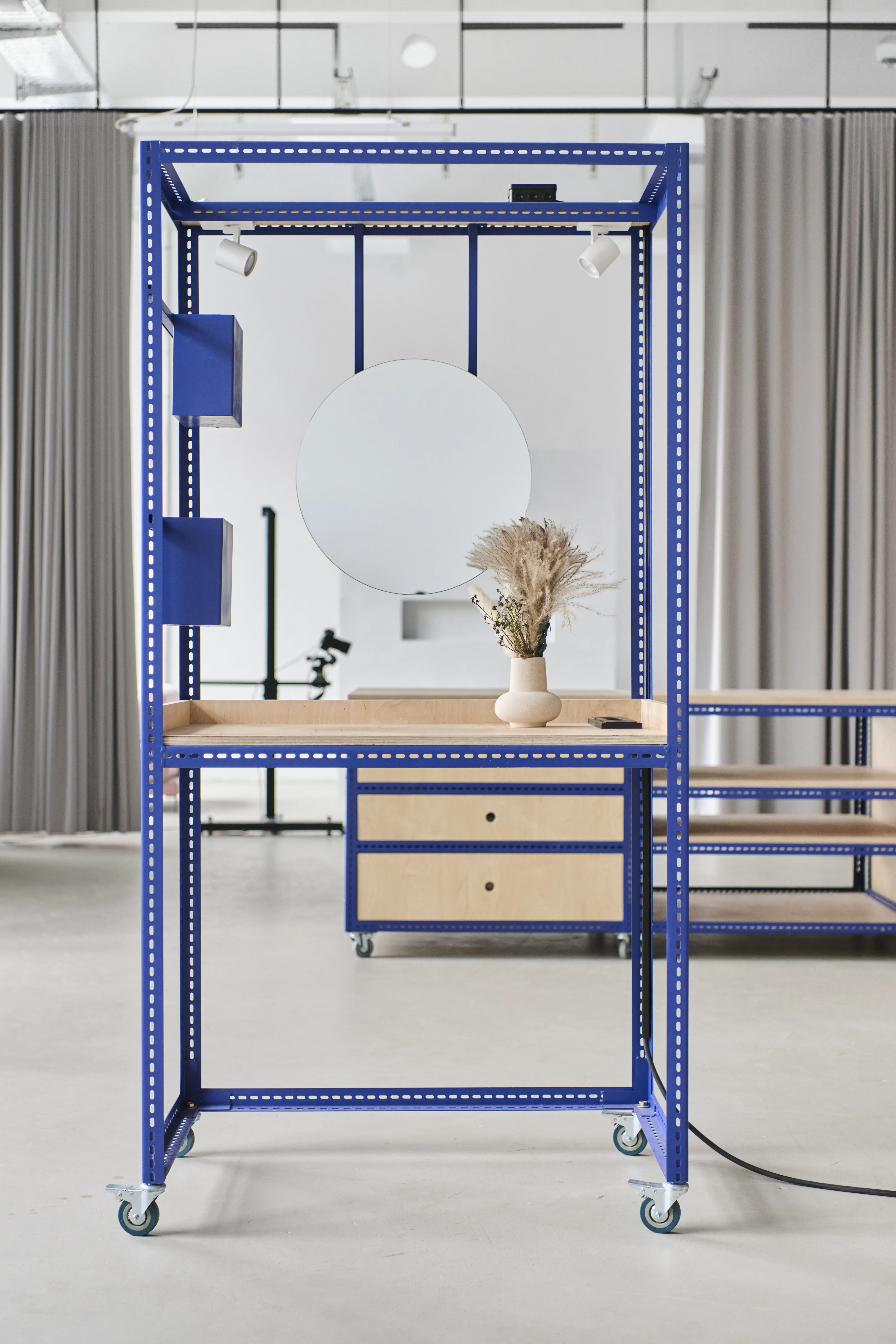 Mobile vanity station at the FLACONI CONTENT STUDIO with a round mirror, spotlights and plywood top in a blue perforated steel frame on castors.