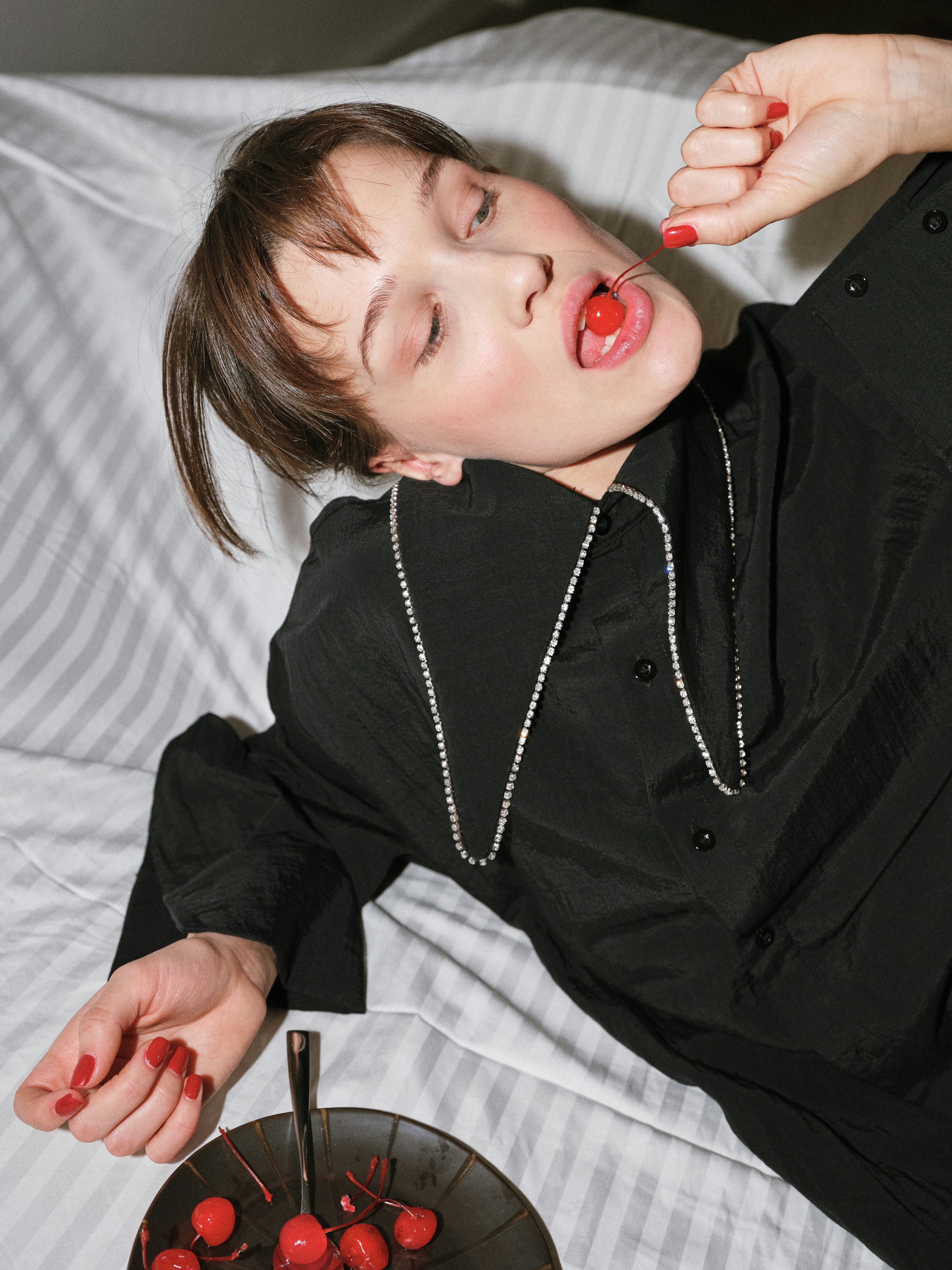 Intimate overhead editorial portrait of a brunette model lying on a striped bed wearing a black shirt and crystal necklace, placing a red cherry at her lips