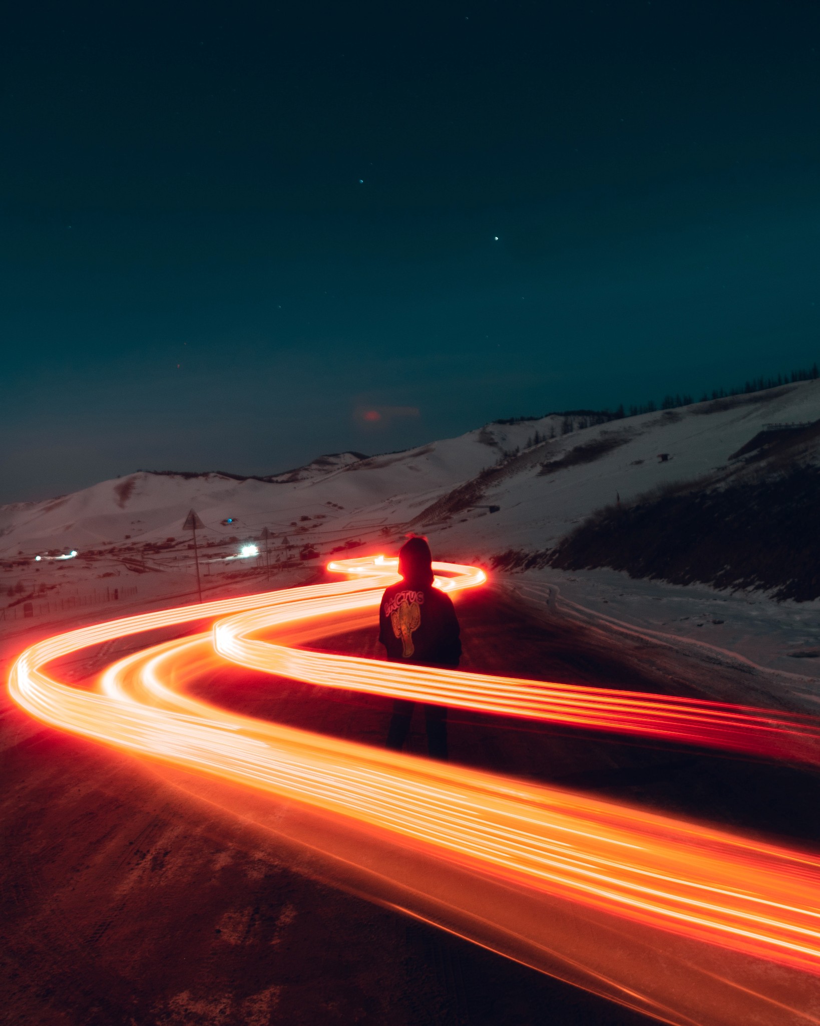 Orange light trails from a car wrapping around a man standing in the road with snowy hills in the background on a clear night