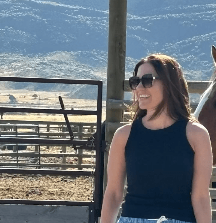 Joanna Mikulski, CEO of Mikulski Strategies LLC, smiling while wearing sunglasses and a black sleeveless top, standing outdoors with a scenic mountain and ranch setting in the background