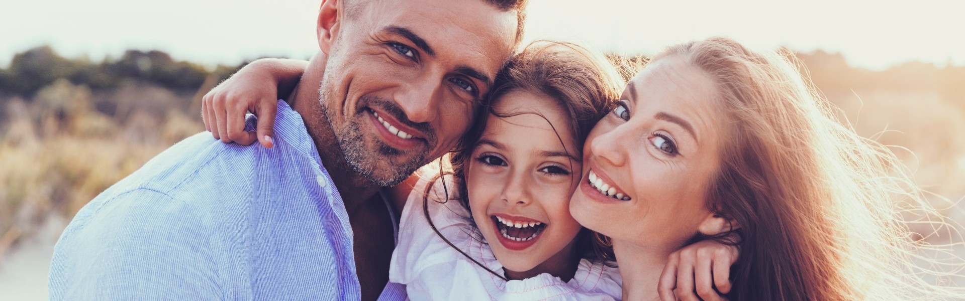 A happy family of three smiling together outdoors at sunset, with a man in a light blue shirt, a young girl in the center, and a woman on the right, all embracing each other closely.