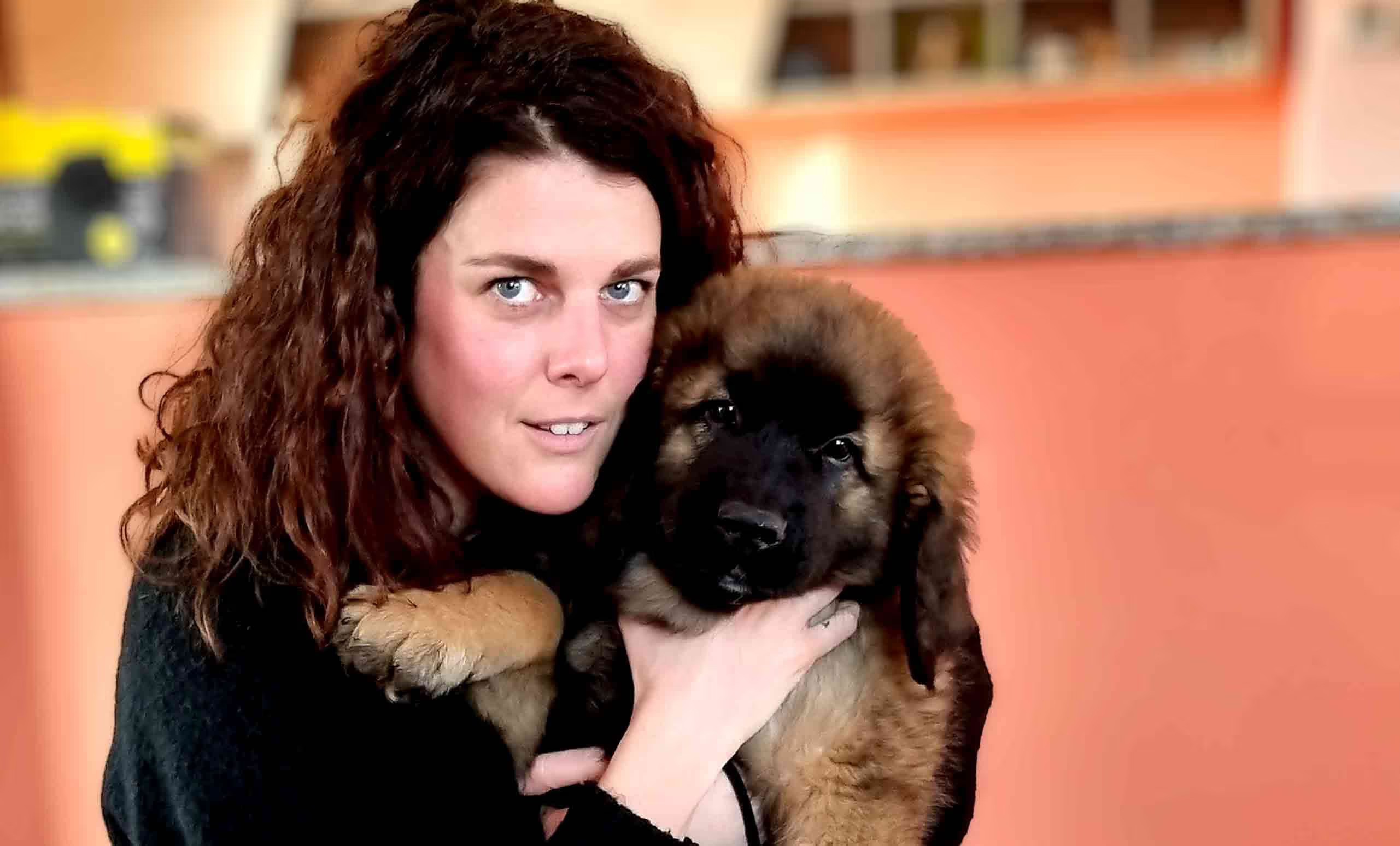 A woman with curly hair smiles while holding a fluffy puppy close to her chest in a cozy indoor setting.