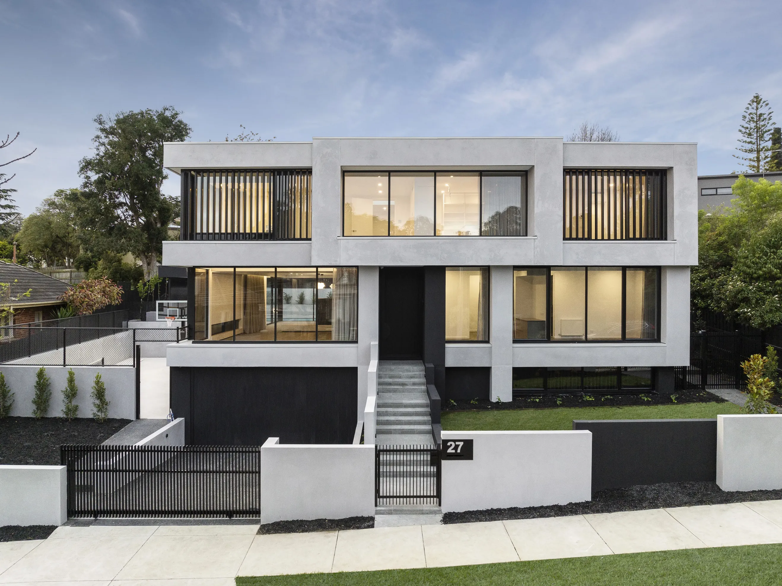 Front facade of Ivanhoe East Residence at dusk, a monochrome two-storey home in textured grey render with black-framed glazing, timber-batten screens, a central stair to a black entry and a black metal fence