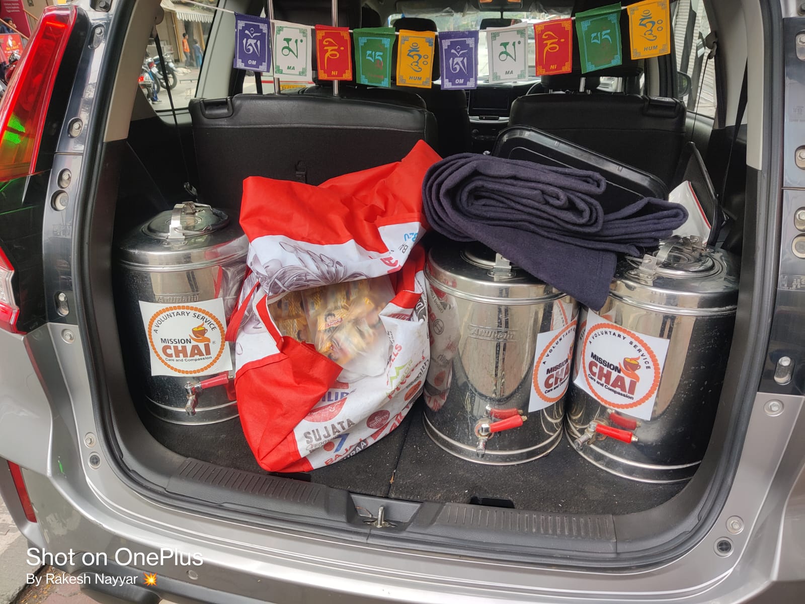 Two volunteers serve tea from a "Mission Chai" dispenser into paper cups on trays, smiling in a hospital corridor.