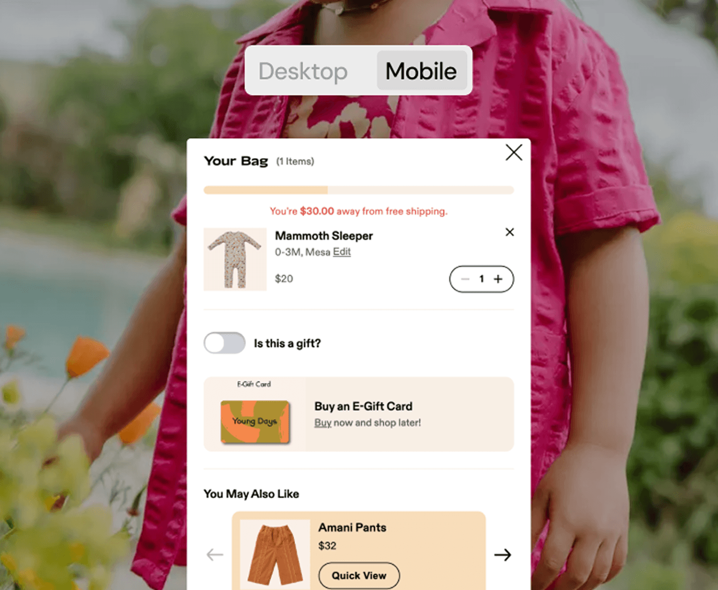 A mobile shopping cart interface is shown over a background image of a child in a bright pink outfit. The cart contains one item labeled “Mammoth Sleeper” priced at $20, with a message indicating the user is $30 away from free shipping. The interface also features gift options, an e-gift card section, and a product suggestion for “Amani Pants.” At the bottom, the subtotal is shown with a “Checkout” button. The view toggle at the top is set to "Mobile."
