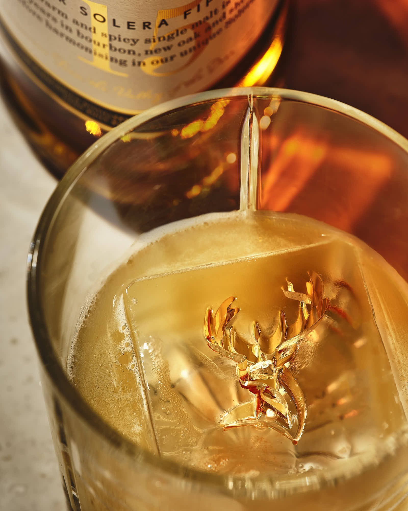 A close-up of a whiskey glass filled with a golden drink, ice cubes, and a bottle in the background.