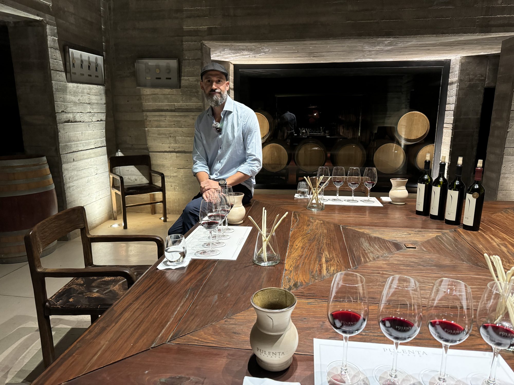 Winemaker Javier Lo Forte seated at a wooden tasting table with red wine glasses and bottles, with wine barrels visible in the Pulenta Estate cellar.