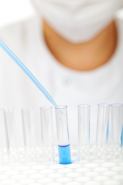 Laborant w maseczce ochronnej pipetuje niebieski płyn do próbówki podczas analizy chemicznej.