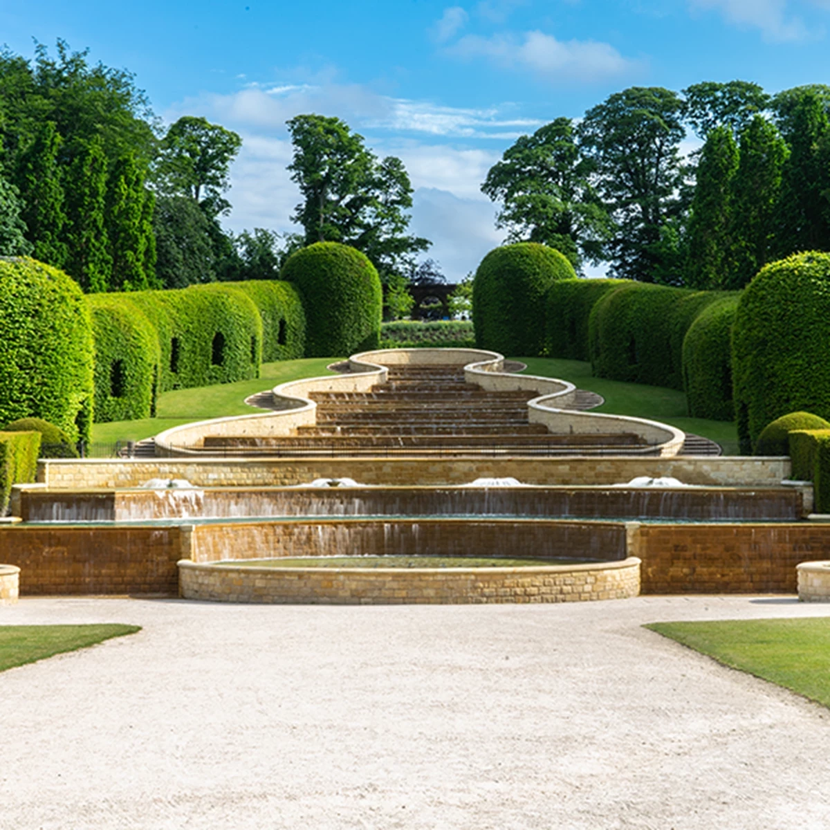 Tiered water feature in a landscaped garden, surrounded by neatly trimmed green hedges and trees under a blue sky.