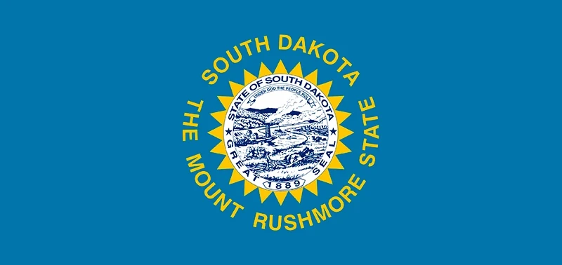 South Dakota state flag featuring a blue field with the state seal in the center. The seal is surrounded by a gold sun design and text: "South Dakota, The Mount Rushmore State."