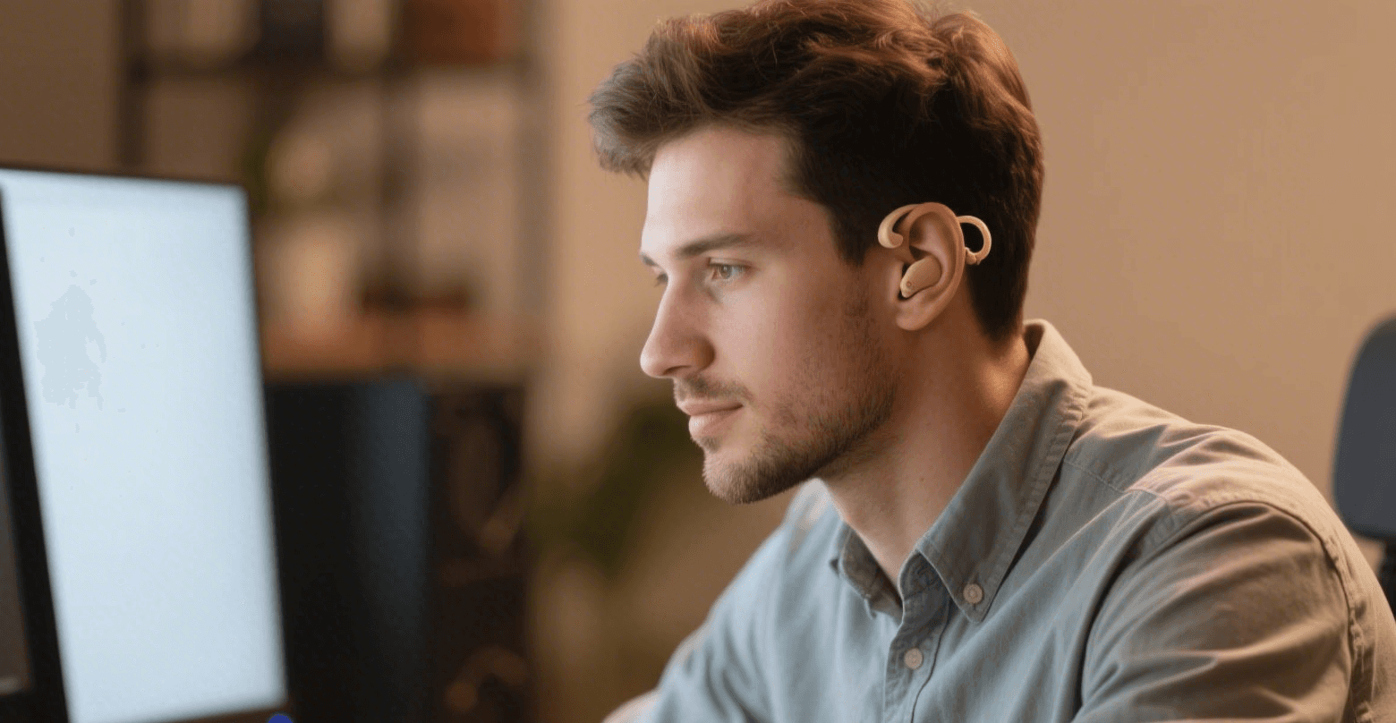 Ein junger Mann mit Bartansatz und einem hellblauen Hemd sitzt vor einem Computermonitor und blickt auf den Bildschirm. Er trägt ein goldenes oder beiges Hörgerät oder Ohrpassstück an seinem rechten Ohr. Der Hintergrund ist unscharf und dunkel, was auf einen Innenraum oder ein Büro hindeutet.