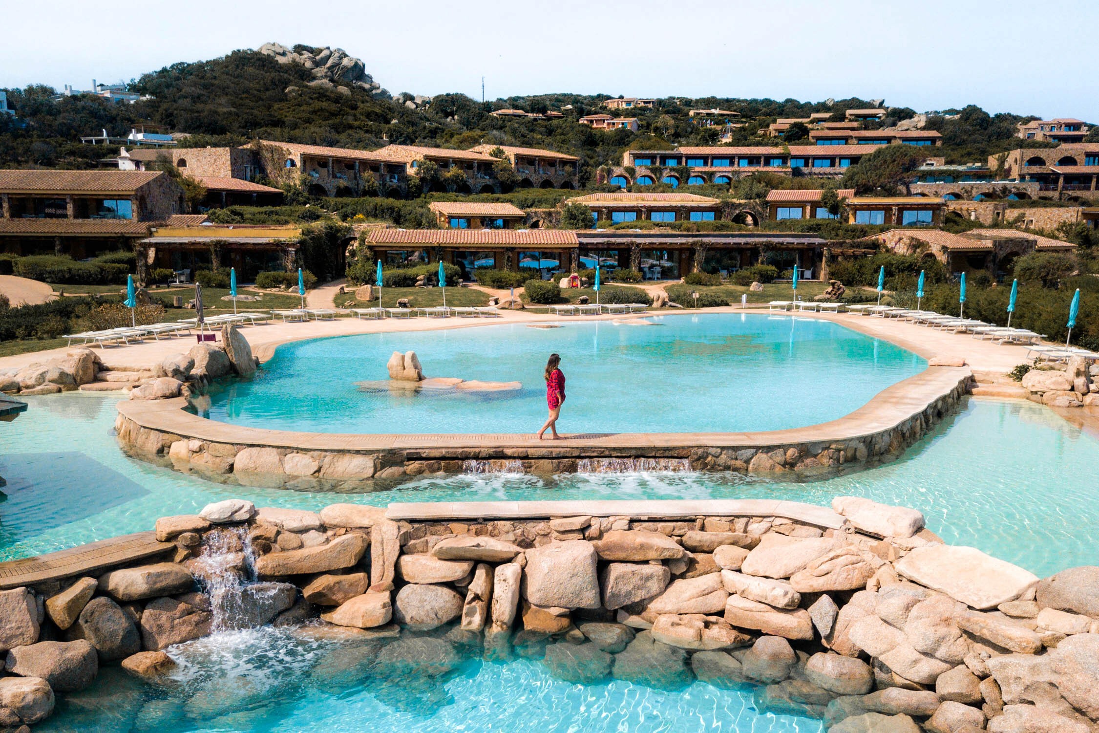 A person in a red swimsuit stands in a turquoise tiered stone pool with waterfalls and resort buildings in the background.