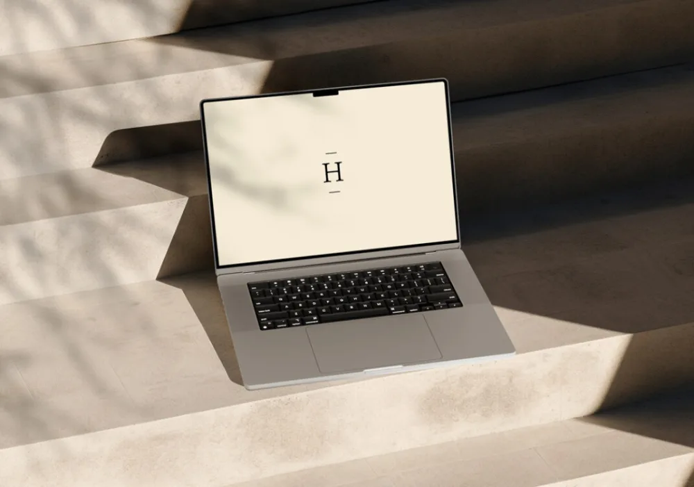 MacBook mockup placed on minimalist concrete steps, bathed in soft natural light with subtle shadows from surrounding foliage. The screen displays a simple, elegant design on a neutral background, emphasizing the laptop's sleek form.