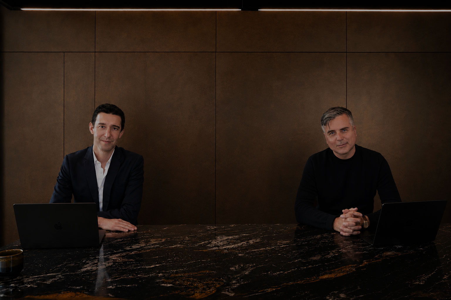 Two people sit at a dark wooden table, smiling warmly at the camera. They have laptops open, creating a friendly and collaborative atmosphere.