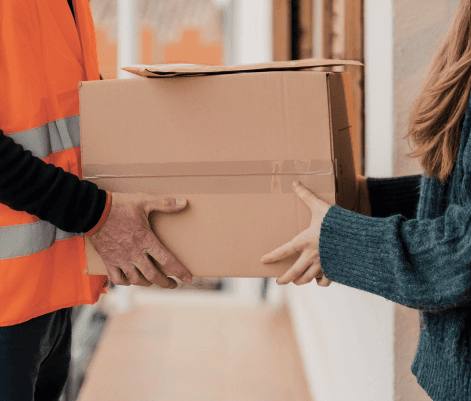 A delivery person in a bright orange vest is handing a cardboard box to a woman standing at a doorway. The woman is wearing a cozy sweater and is reaching out to receive the package. The scene captures a moment of delivery in a residential setting.