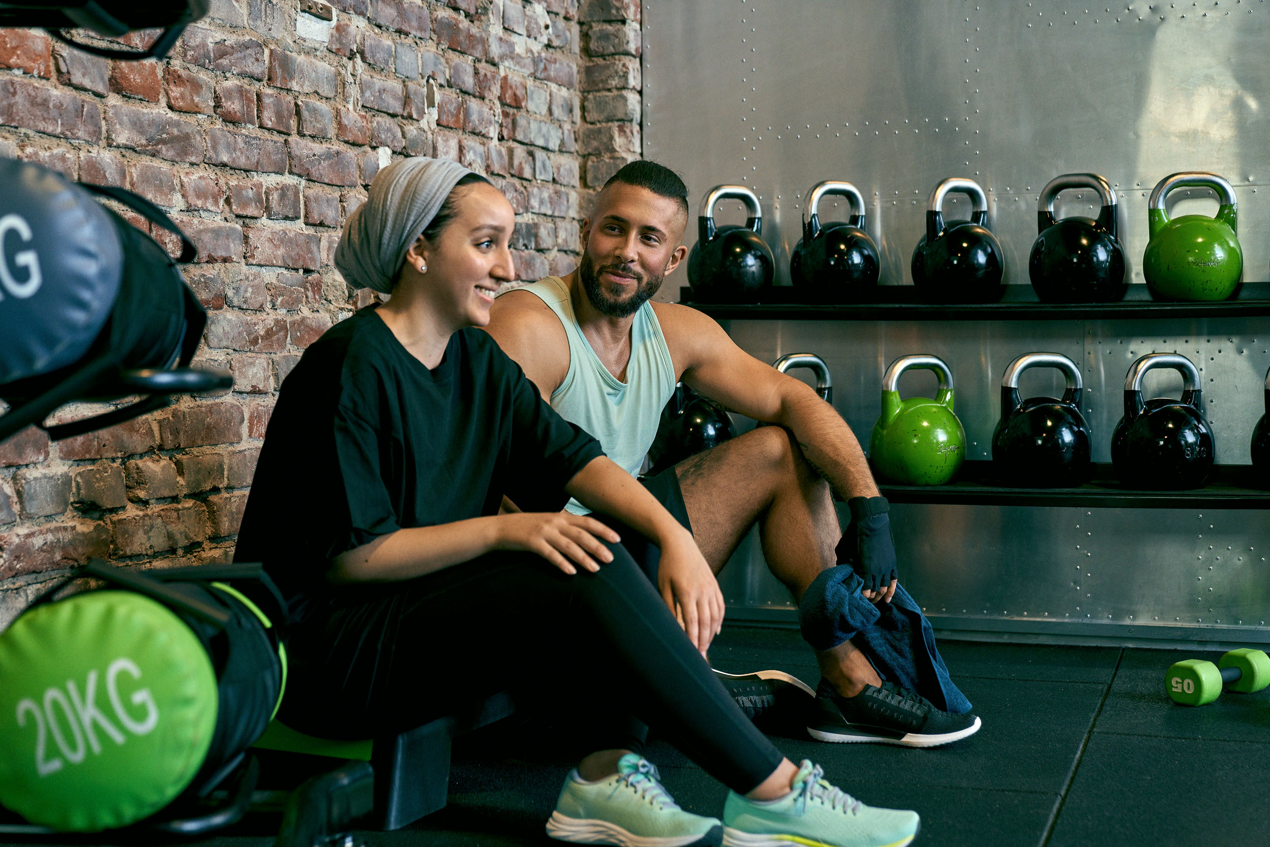 Zwei glückliche Fitnessstudio-Mitglieder entspannen sich beim Gespräch im XTRAFIT Gym, umgeben von farbigen Kettlebells. Die Umgebung im XTRAFIT Gym ist modern, hell und die Geräte sind innovativ und neu.