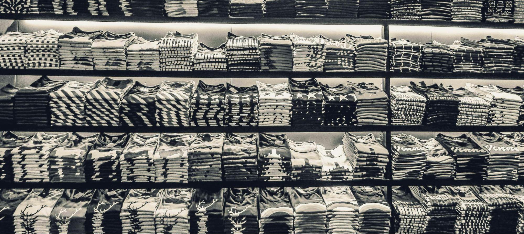 A neatly arranged display of folded shirts on multiple shelves. The shirts come in various patterns and designs, including stripes, polka dots, and abstract prints. The colors range from black and white to more vibrant hues. The shirts are neatly folded and stacked, creating a visually appealing and organized display.