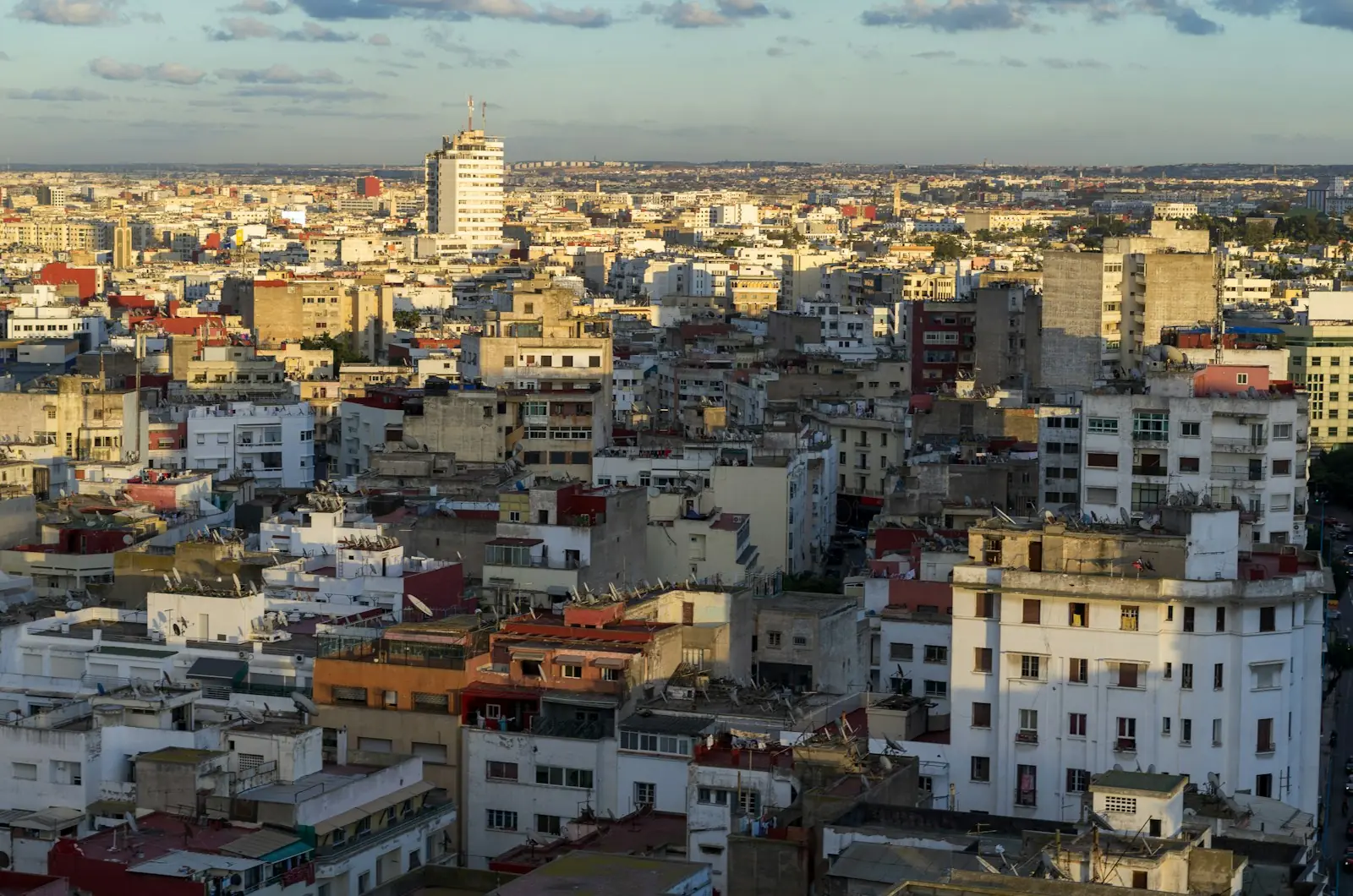 Aerial view of dense white apartment blocks and rooftops across Casablanca in evening light, with a tall tower rising on the horizon