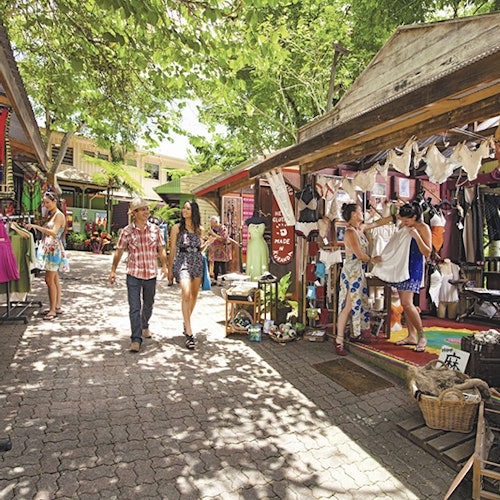 在树木环绕的遮阳摊位下，人们正在户外市场选购服装和配饰。