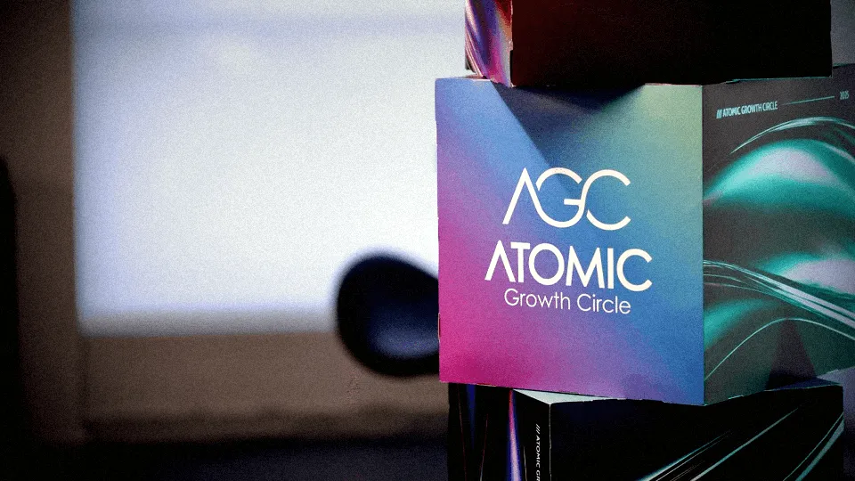 A close-up of a colorful card displaying the text "AGC ATOMIC GROWTH CLUB" against a blurred background. Atomic Growth Club. Atomic Group. Br24. 